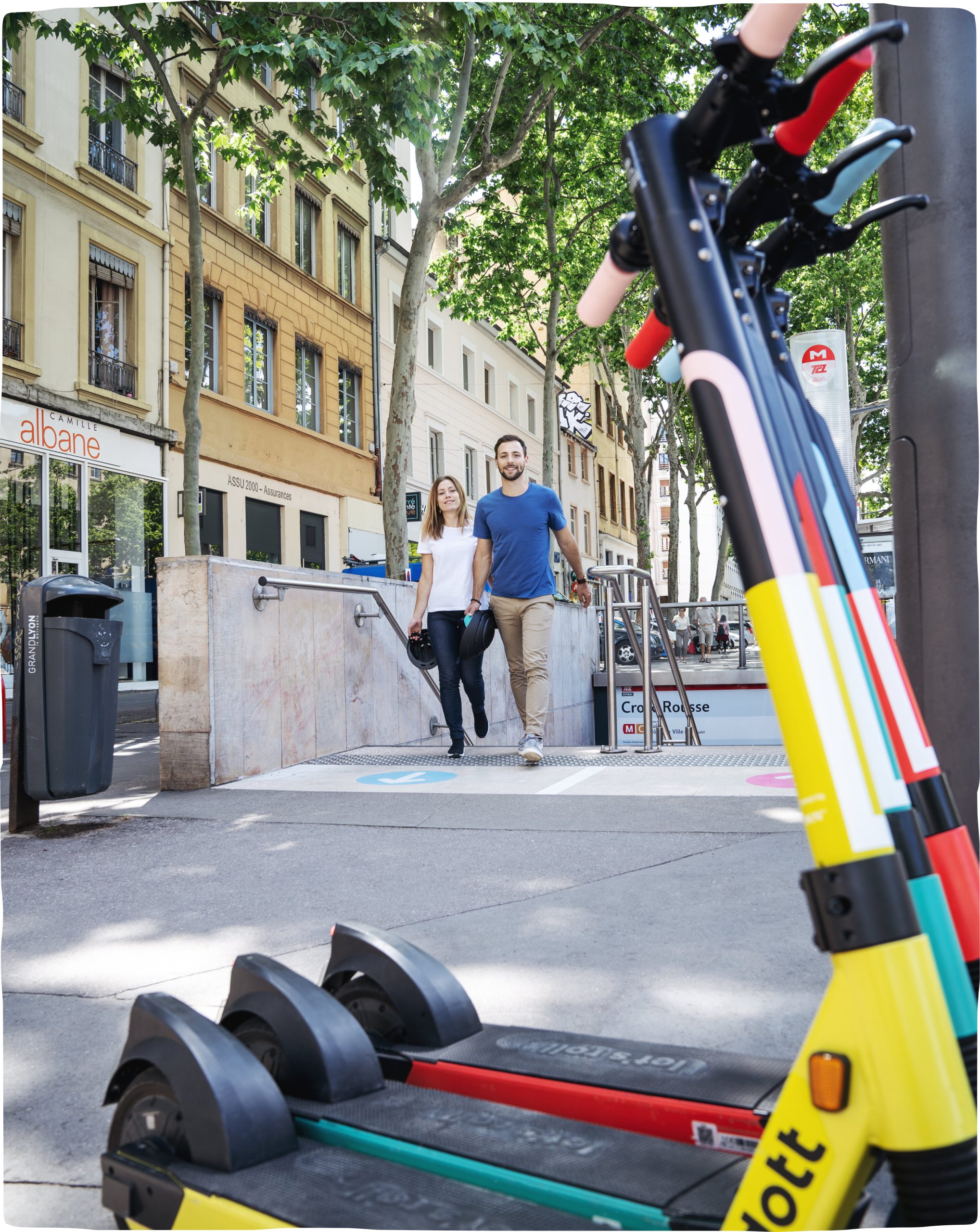 A man and a woman walking out of a metro station carrying helmets to find Dott scooters parked on the side in front of them, ready to ride.
