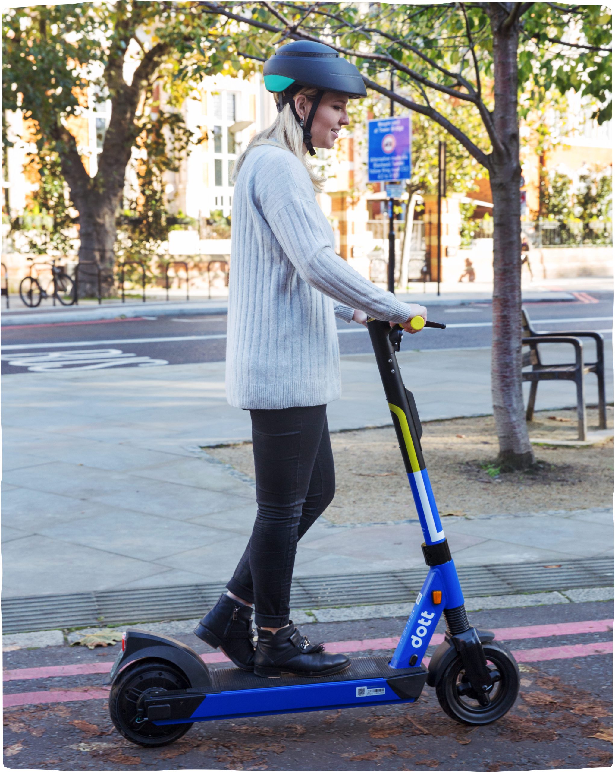 A woman rides a blue Dott through the streets of London wearing a helmet. Sunlight reflects off of buildings in the background.