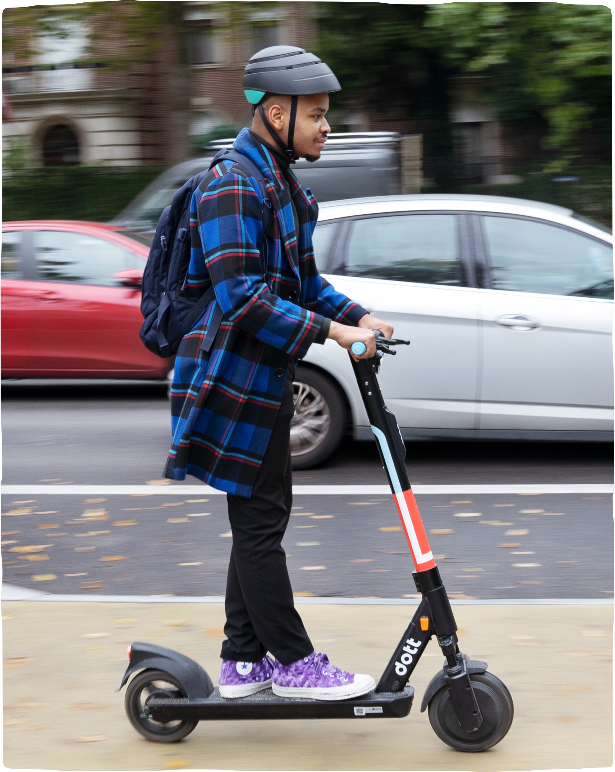 A young man rides a Dott along a city street. He wears a helmet. The cars in the background are blurry, implying he is in motion.