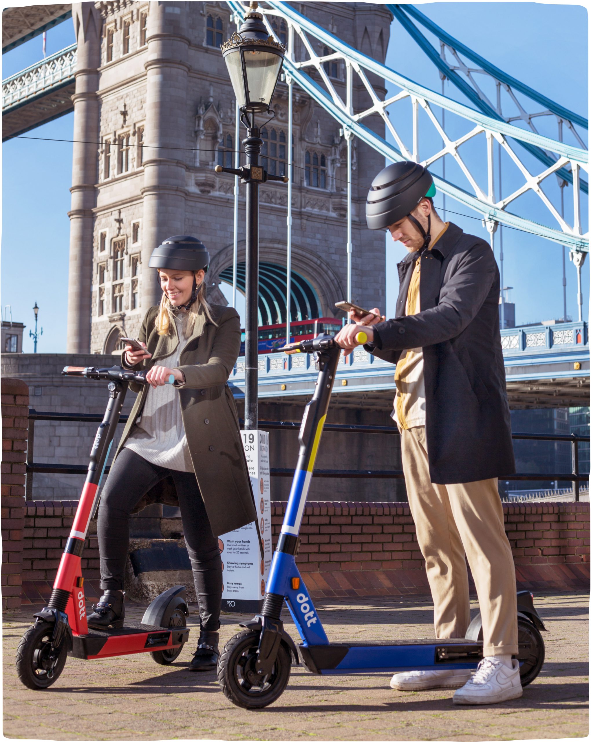 A woman and man each wearing helmets and standing with an e-scooter, while holding their phones to scan the e-scooter and park in designated areas in the city.