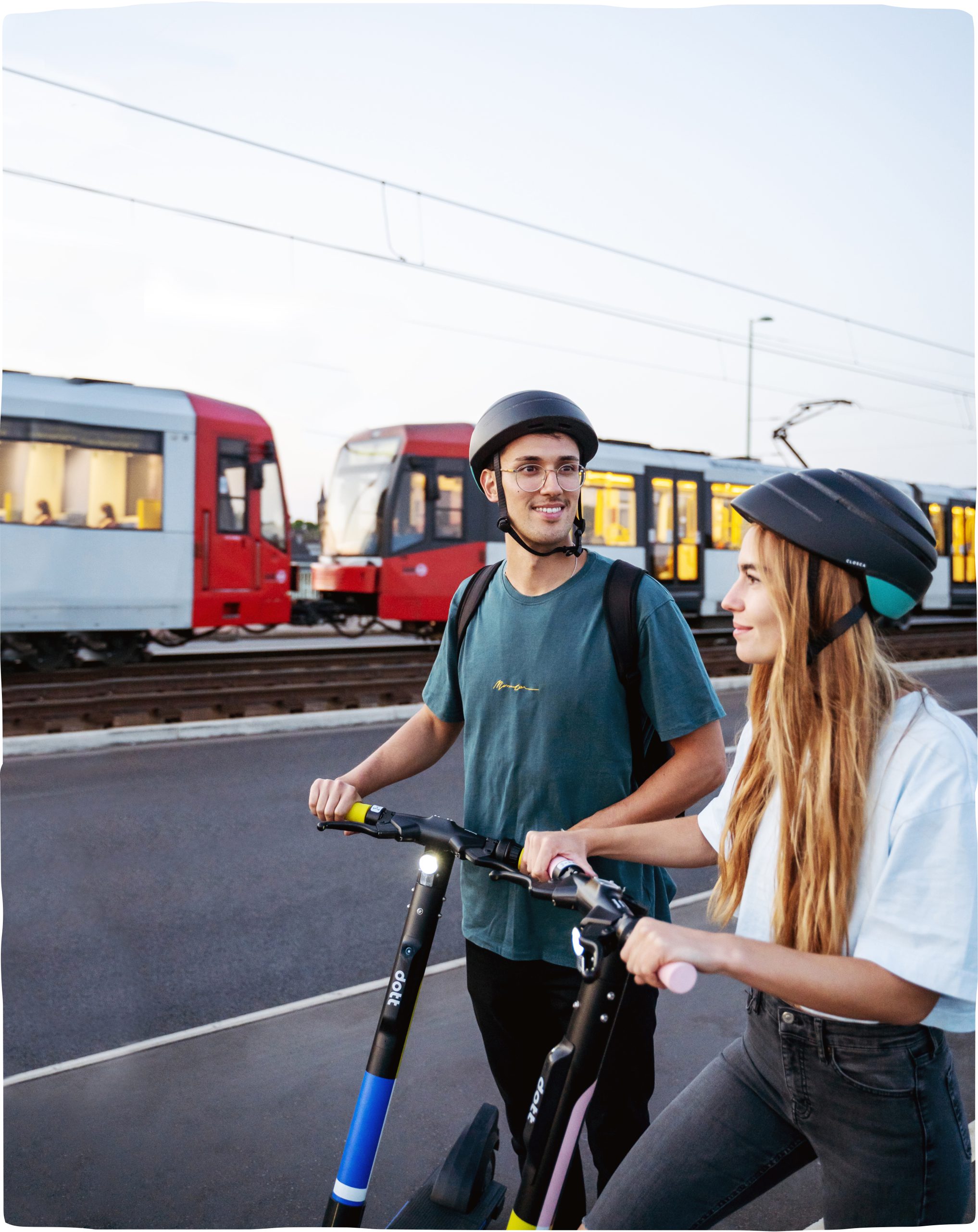 A picture of a woman and a man riding Dott scooters next to each other standing next to a train going past.