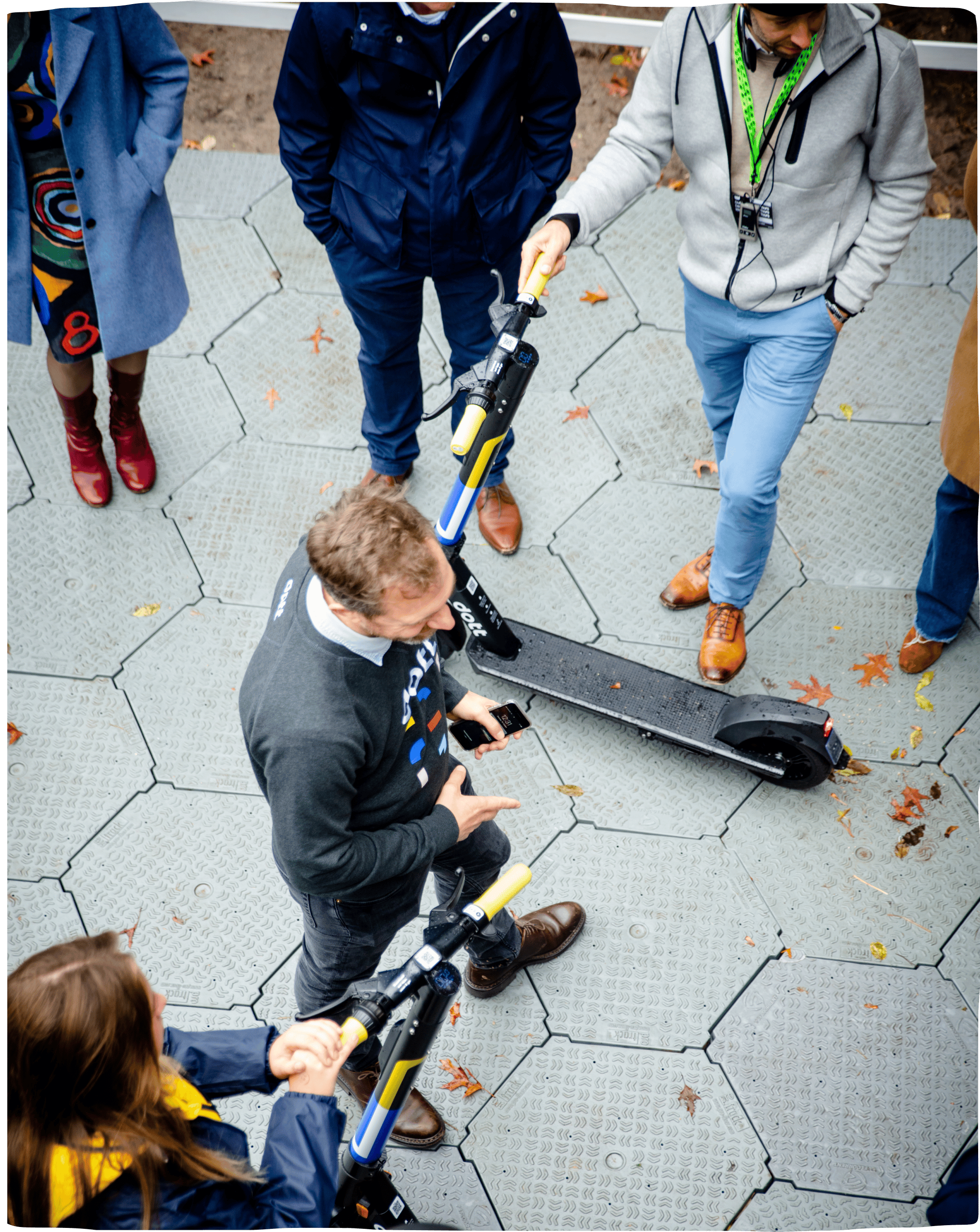 A photo shot from above with a man in the middle, surrounded by people listening to him speak about Dott, with Dott e-scooters on each side.