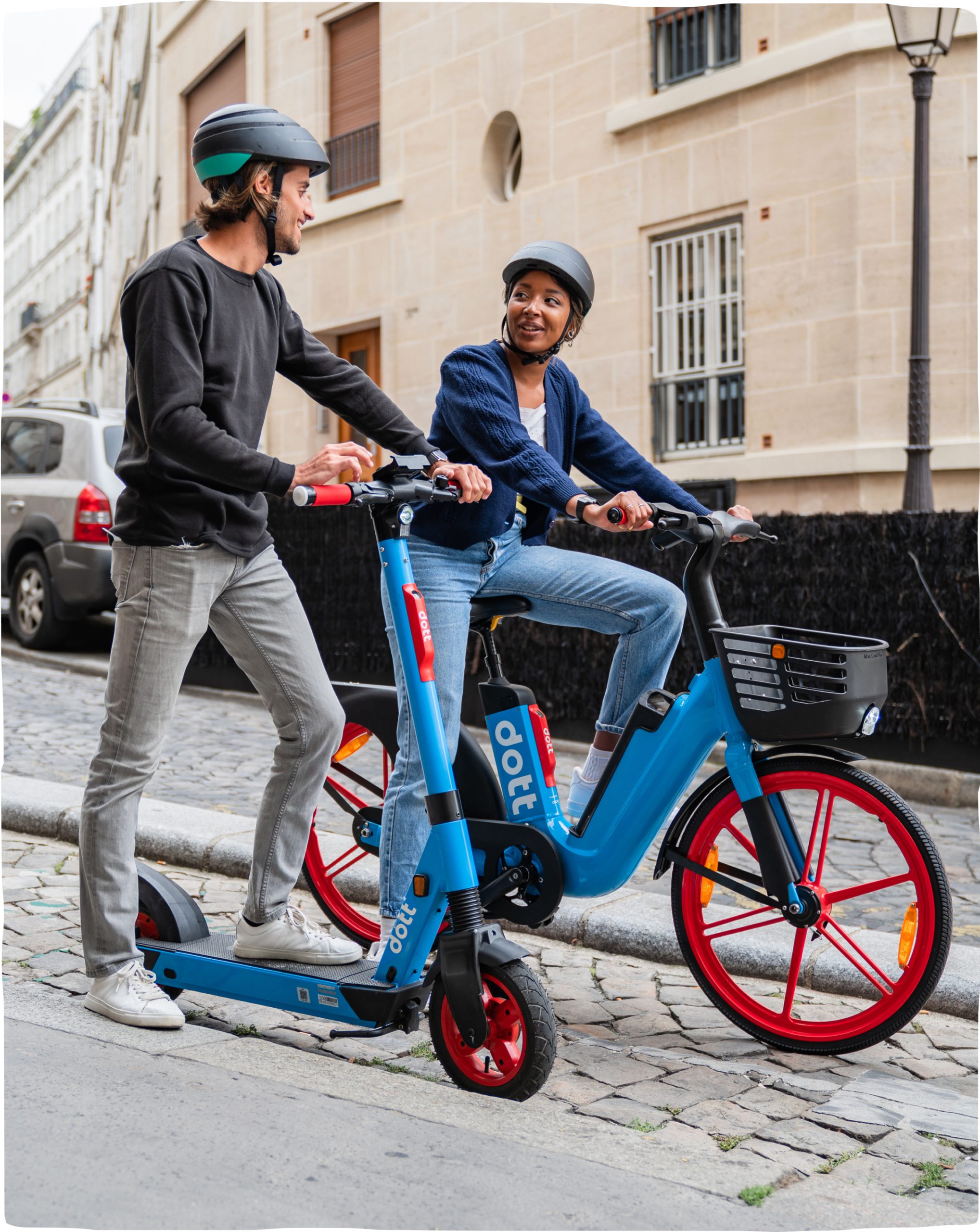 A man on a Dott e-scooter and a woman on a Dott e-bike stand on the European cobblestone sidewalk to talk. They’re both wearing Dott helmets and are smiling at each other.