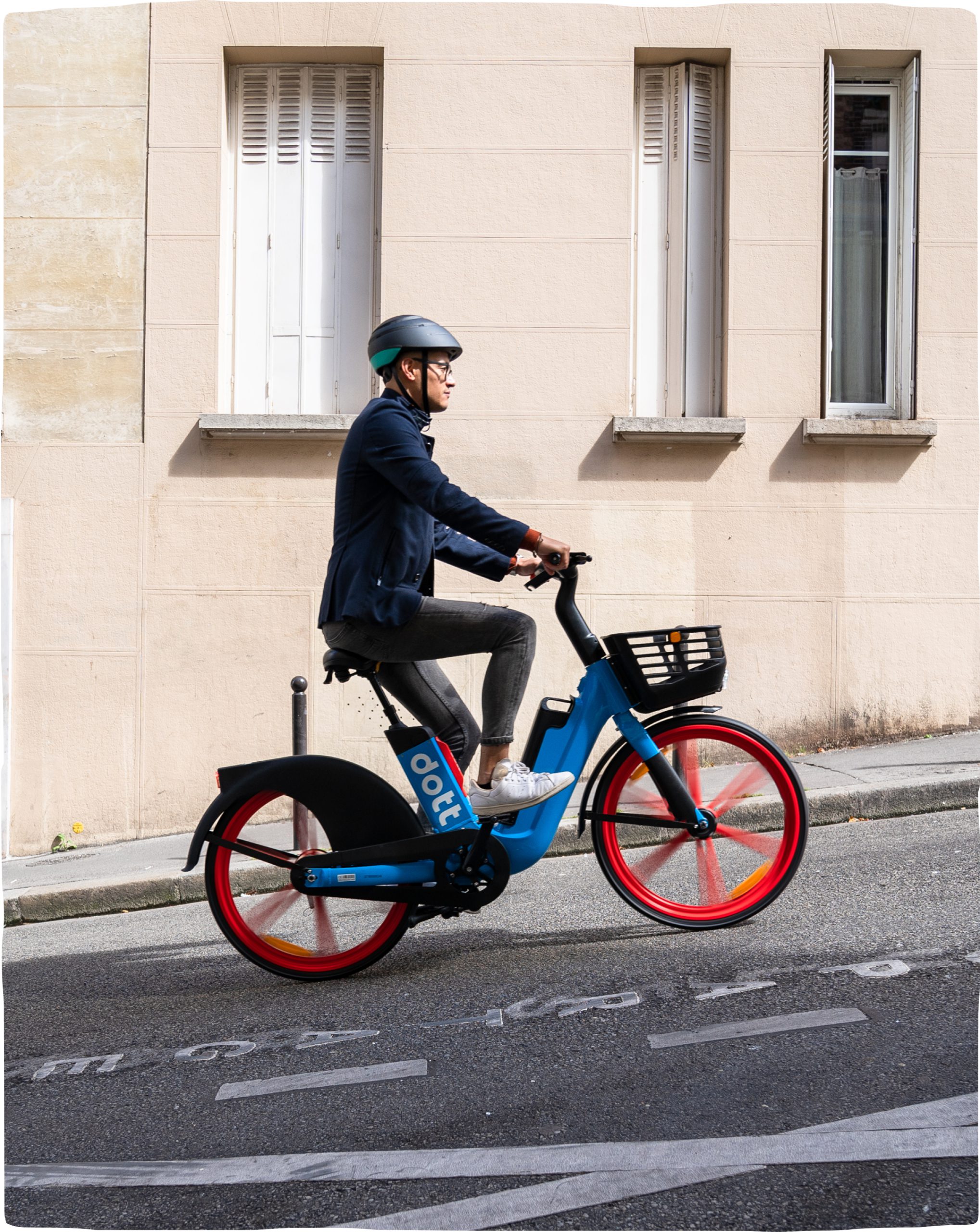 An image of a man riding the Dott e-bike up a hill. The wheels are blurred to indicate movement. The sun shines on the building in the background.