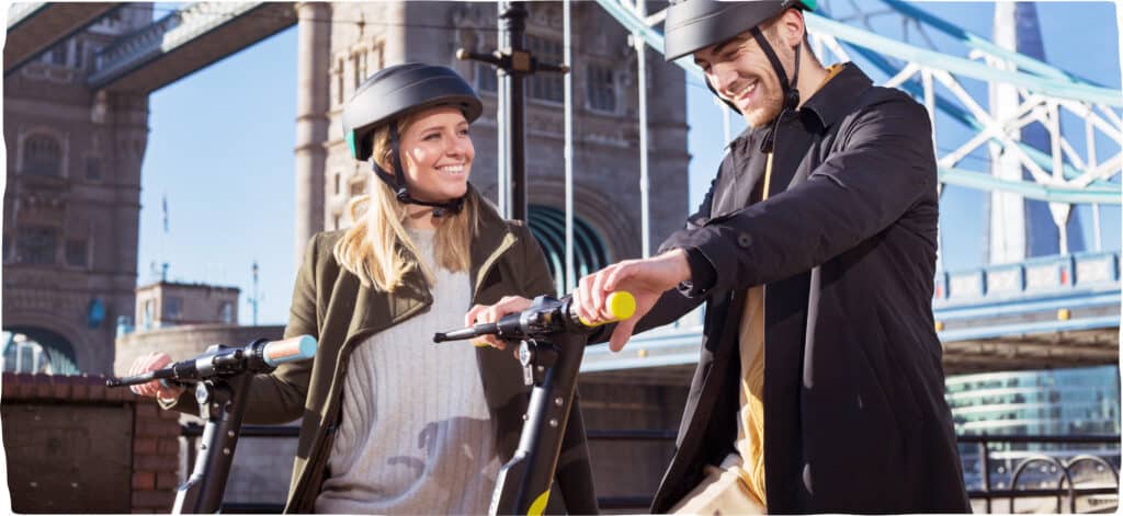 A man and a woman are wearing helmets and smiling. The woman is turned to face the man whilst the man has both hands on his Dott handlebars.