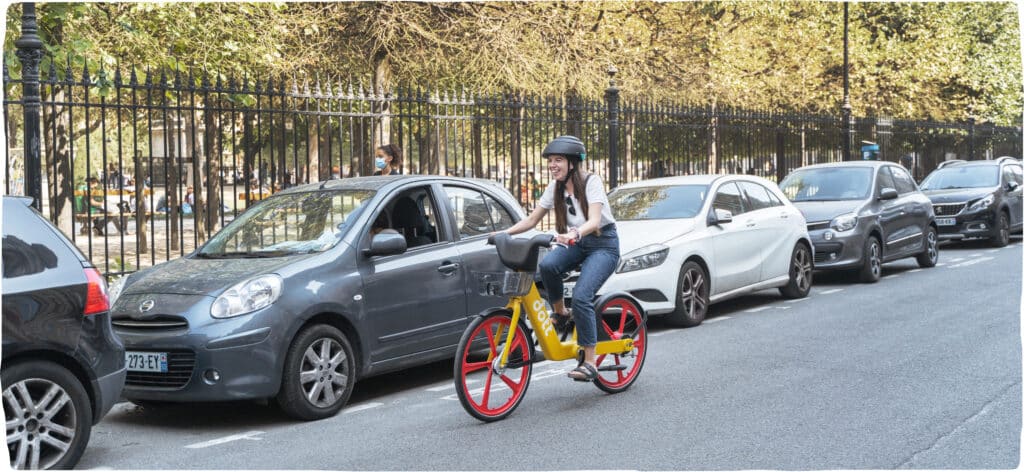 A woman wearing a helmet, smiling and riding a yellow e-bike along a park with a row of cars parked behind her.