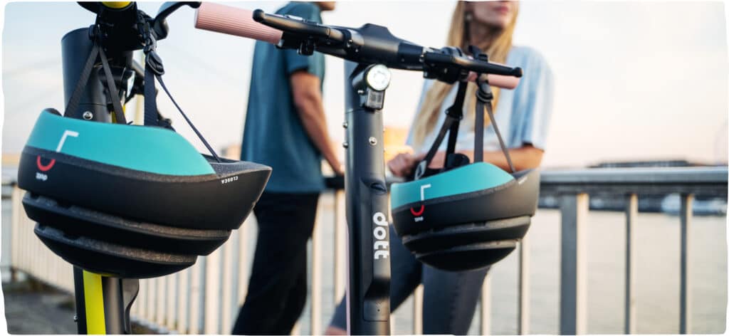 Two Dott helmets hanging on parked scooters next to two people stopped on the side of a bridge enjoying the scenery.