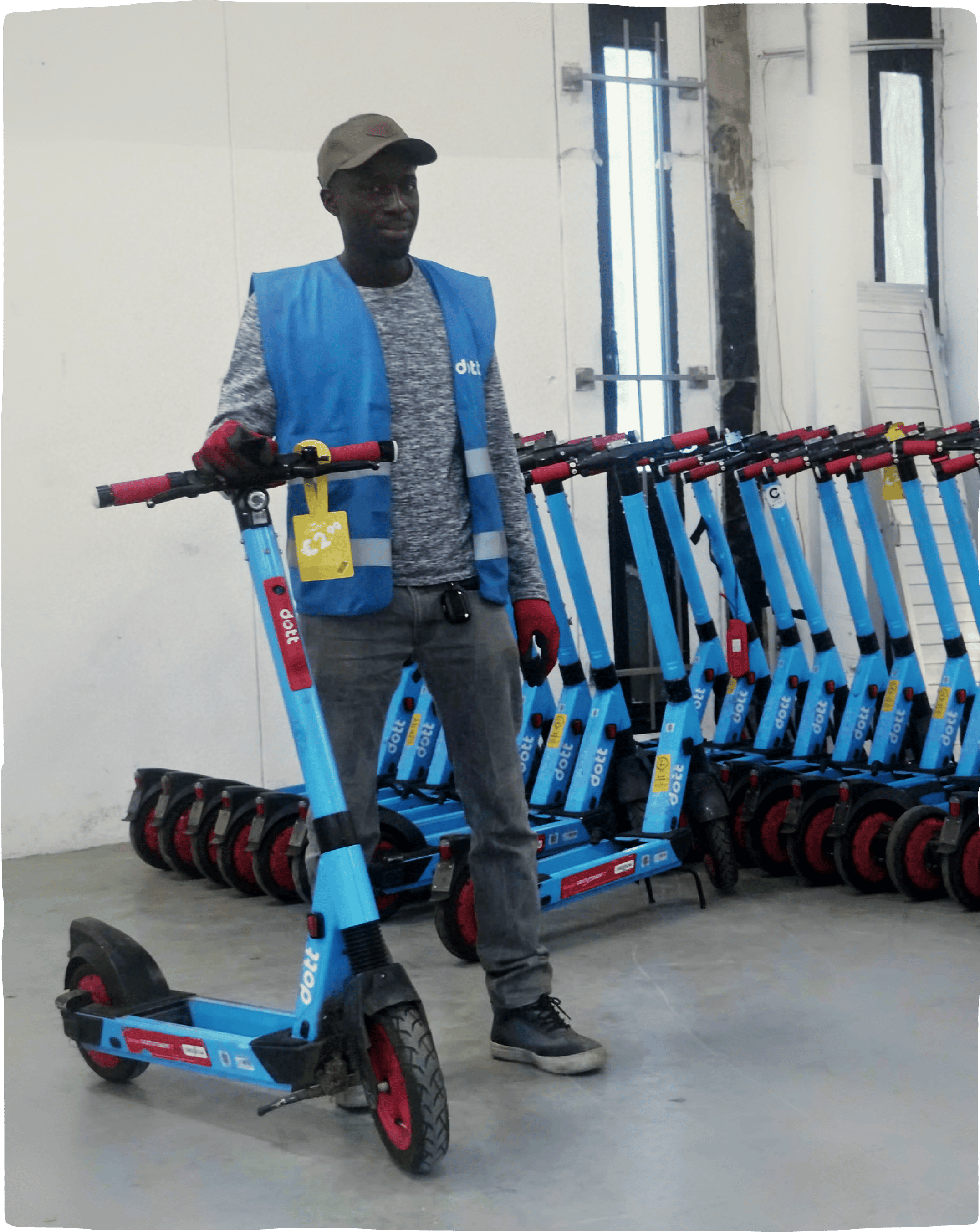An image of a man standing with a Dott e-scooter inside the Dott warehouse, with a row of parked e-scooters in the background.